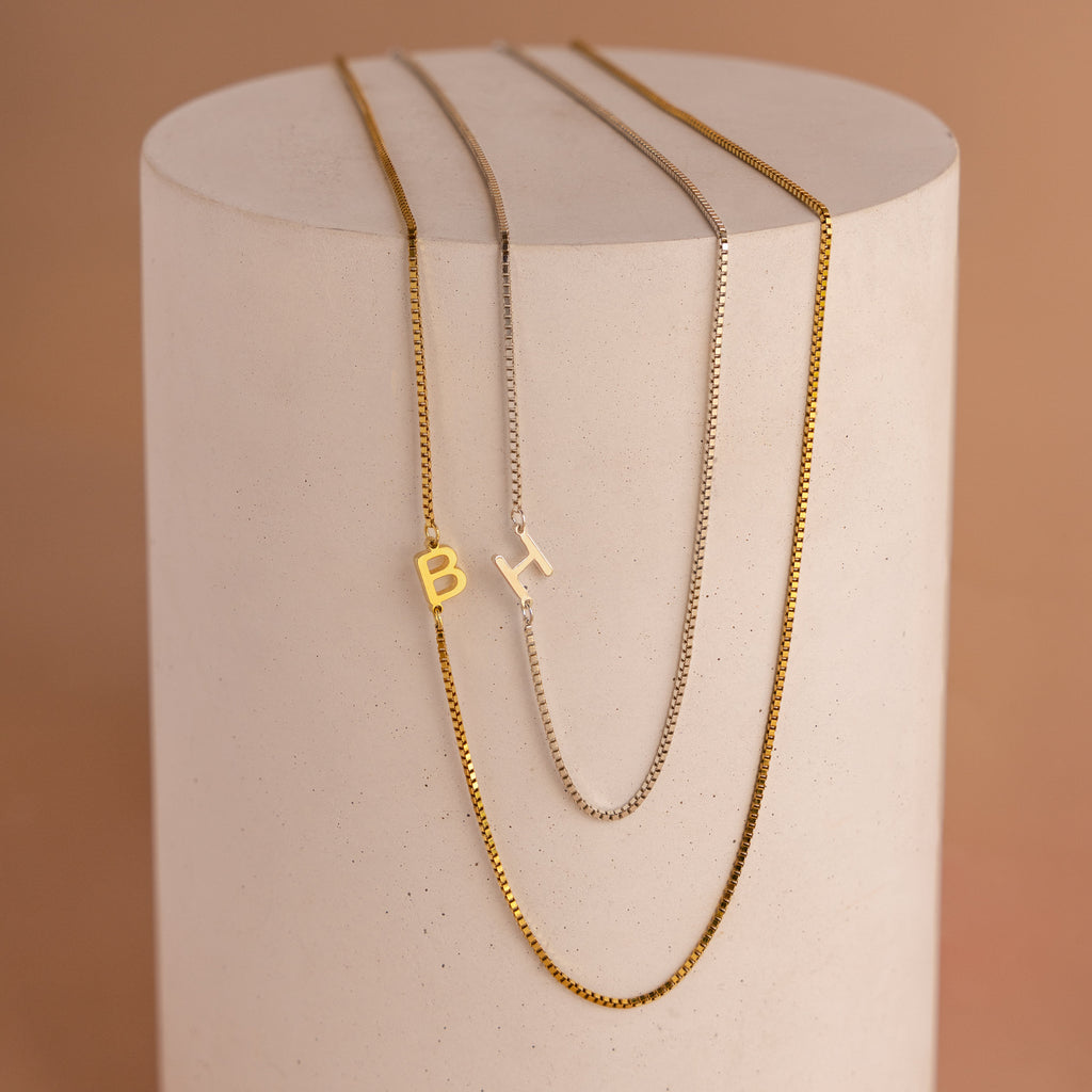 Two gold Sideways Initial Necklaces in box chain and one silver with letter pendants B and H, displayed on a cylinder—perfect for adding an initial touch to any style.