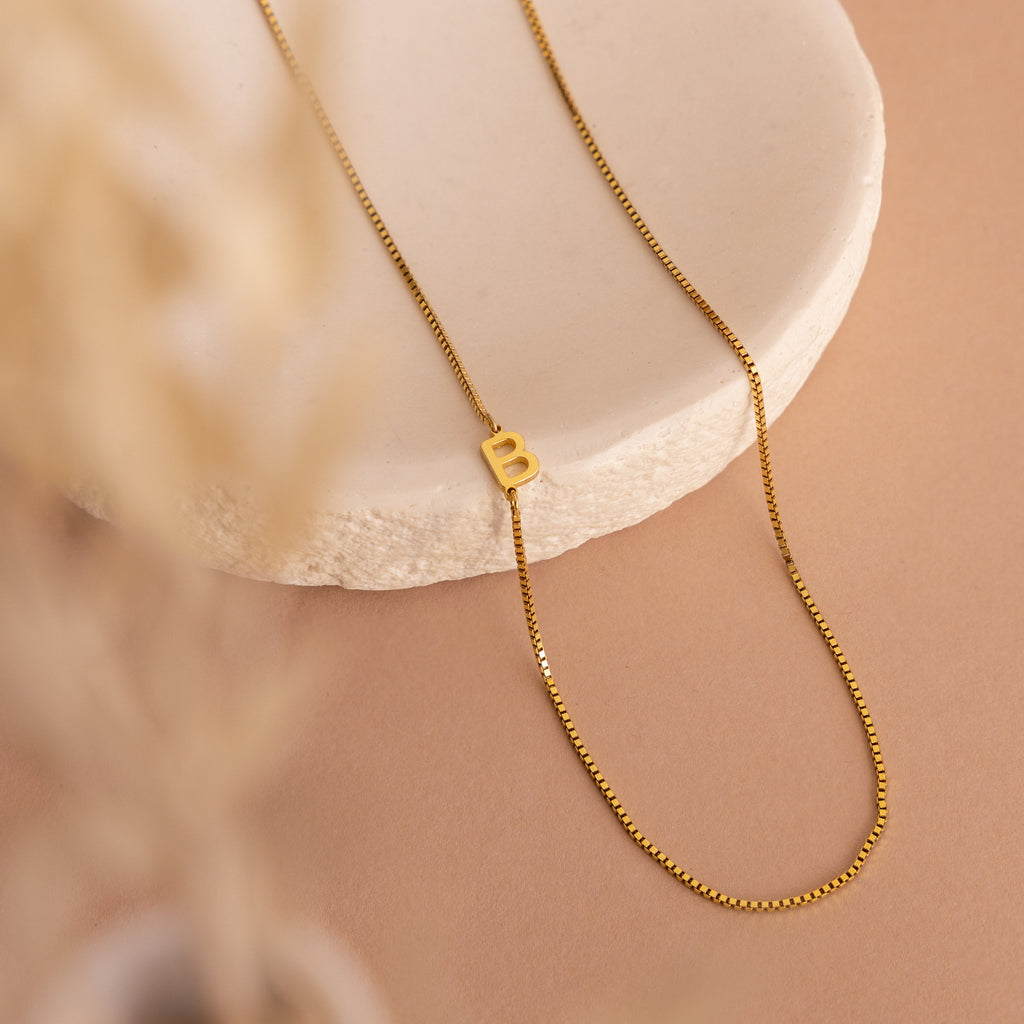 The Sideways Initial Necklace In Box Chain with a “B” pendant is displayed on a round white stone slab against a beige background—ideal for anyone who loves personalized initial jewelry.