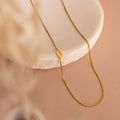 The Sideways Initial Necklace In Box Chain with a “B” pendant is displayed on a round white stone slab against a beige background—ideal for anyone who loves personalized initial jewelry.