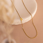 The Sideways Initial Necklace In Box Chain with a “B” pendant is displayed on a round white stone slab against a beige background—ideal for anyone who loves personalized initial jewelry.