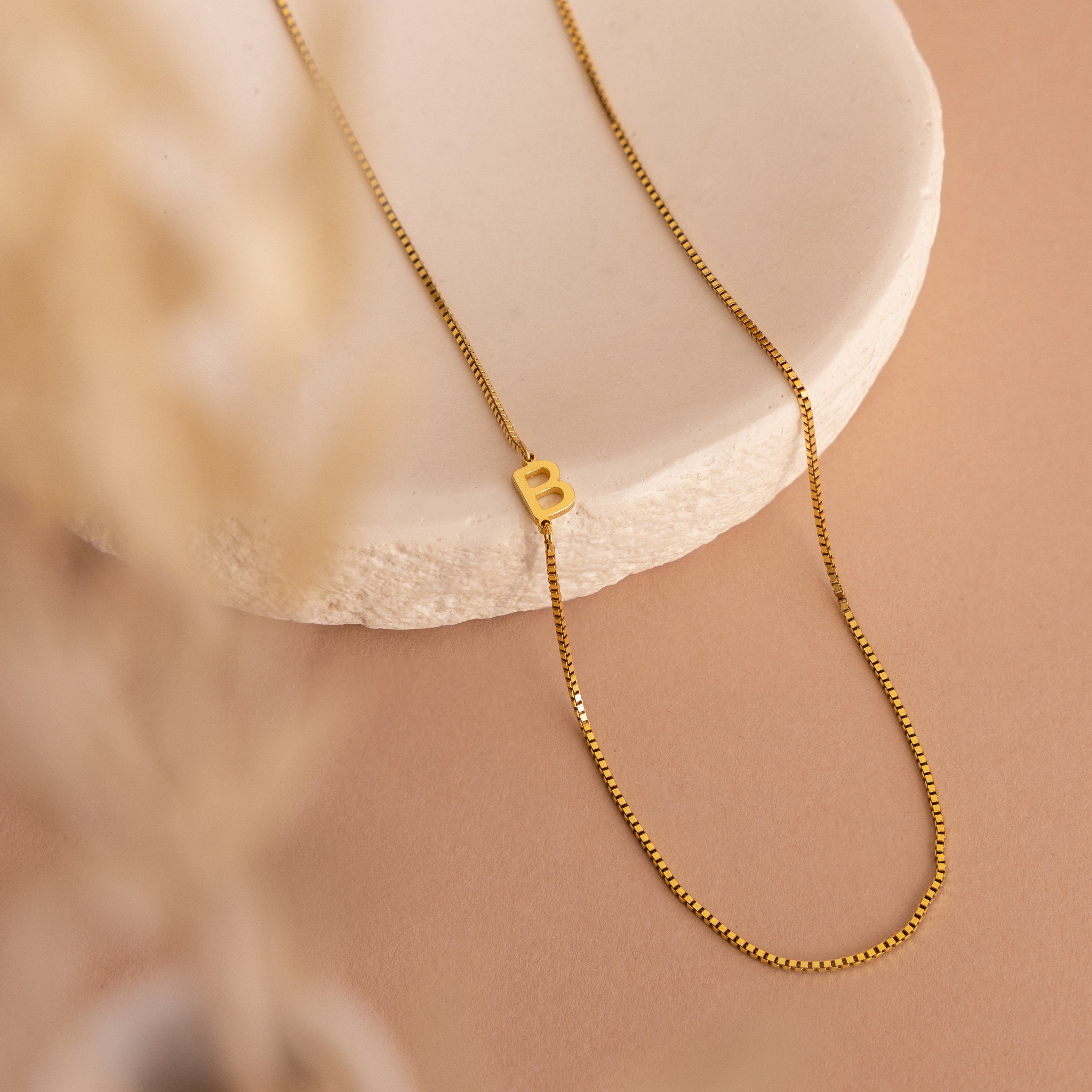 The Sideways Initial Necklace In Box Chain with a “B” pendant is displayed on a round white stone slab against a beige background—ideal for anyone who loves personalized initial jewelry.