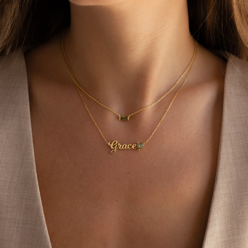 Close-up of a woman wearing two gold necklaces, including the Mellow Name Heart Birthstone Necklace featuring "Grace" and a small heart charm.