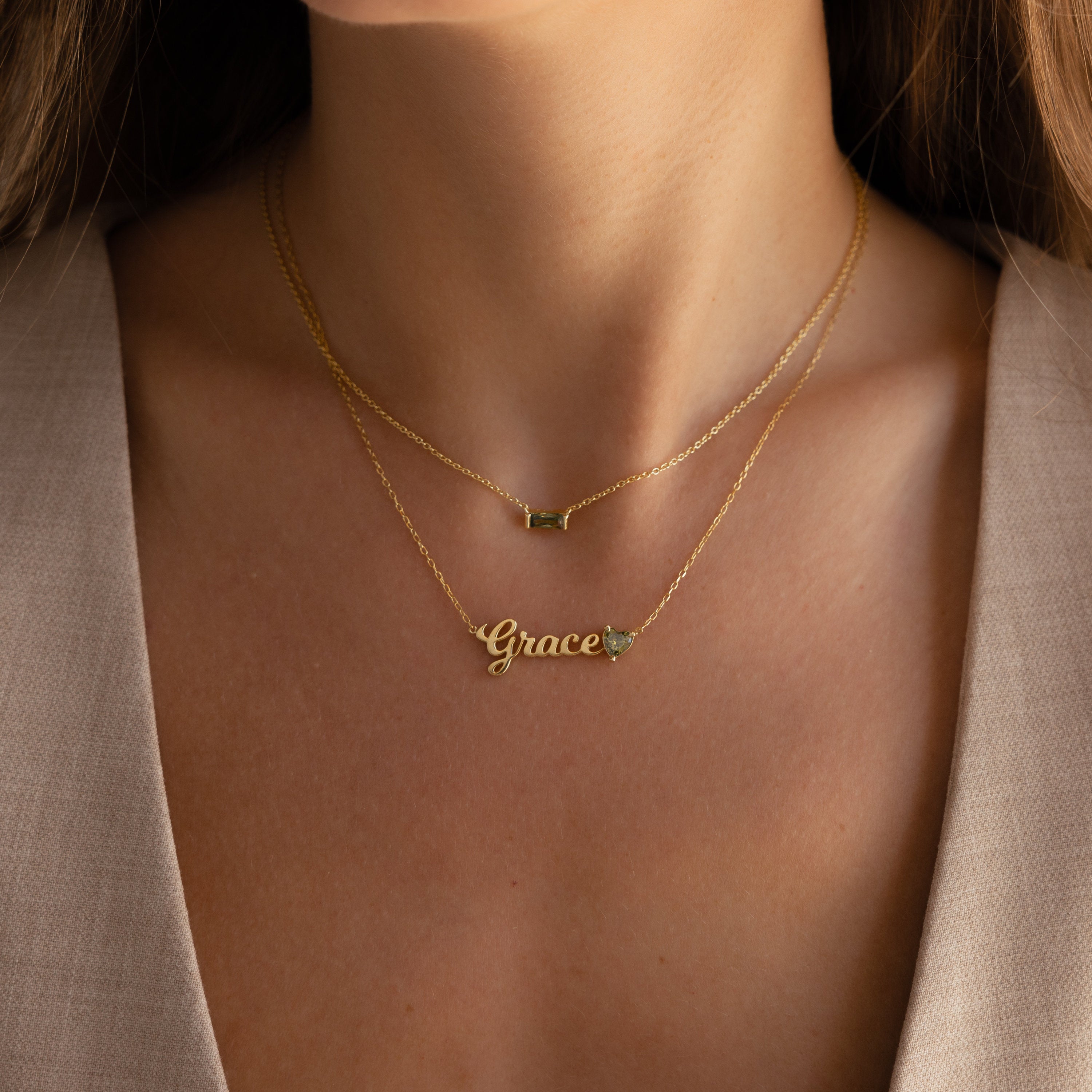 Close-up of a woman wearing two gold necklaces, including the Mellow Name Heart Birthstone Necklace featuring "Grace" and a small heart charm.
