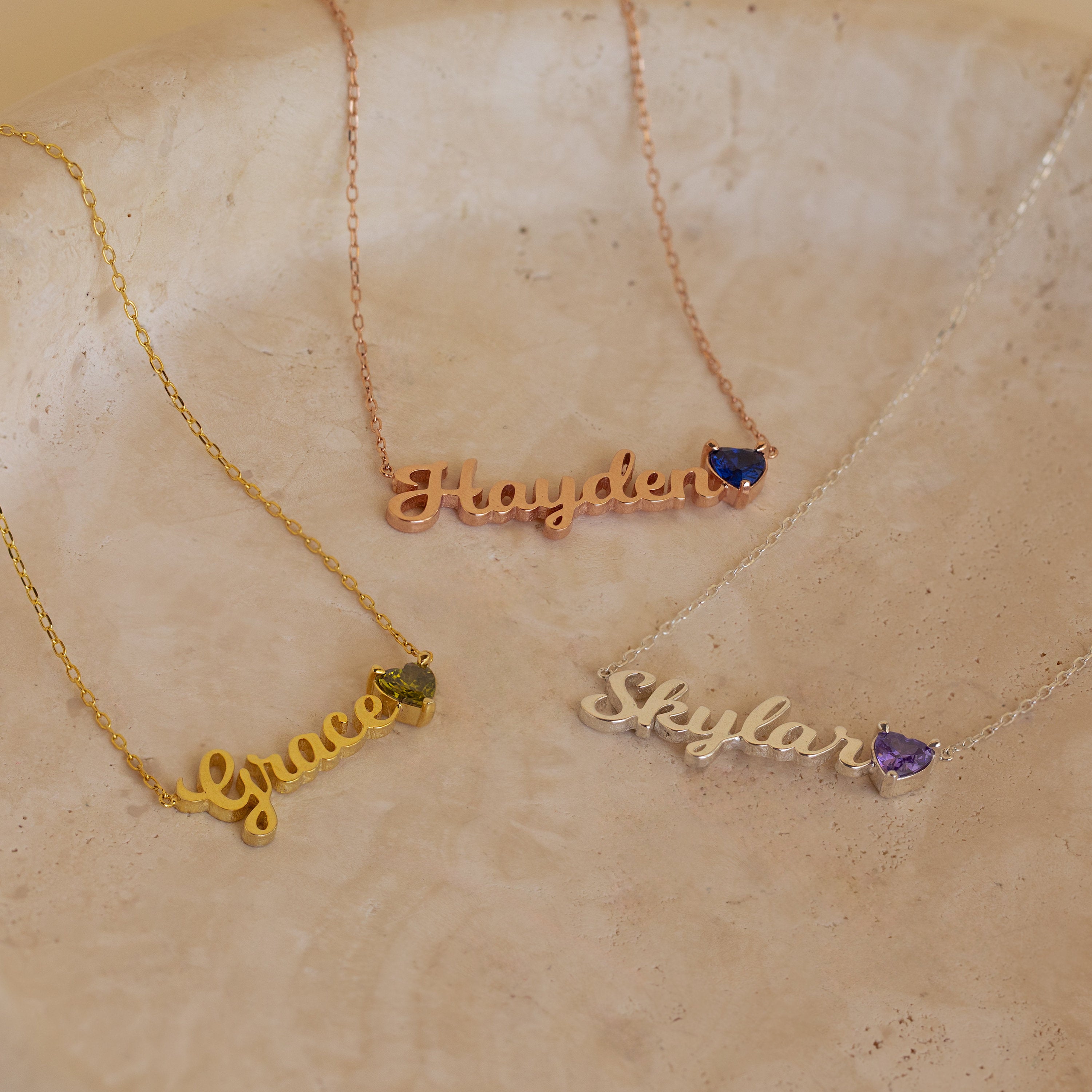 Three Mellow Name Heart Birthstone Necklaces labeled Grace, Hayden, and Skylar, each with a delicate birthstone gem, displayed on a marble surface.
