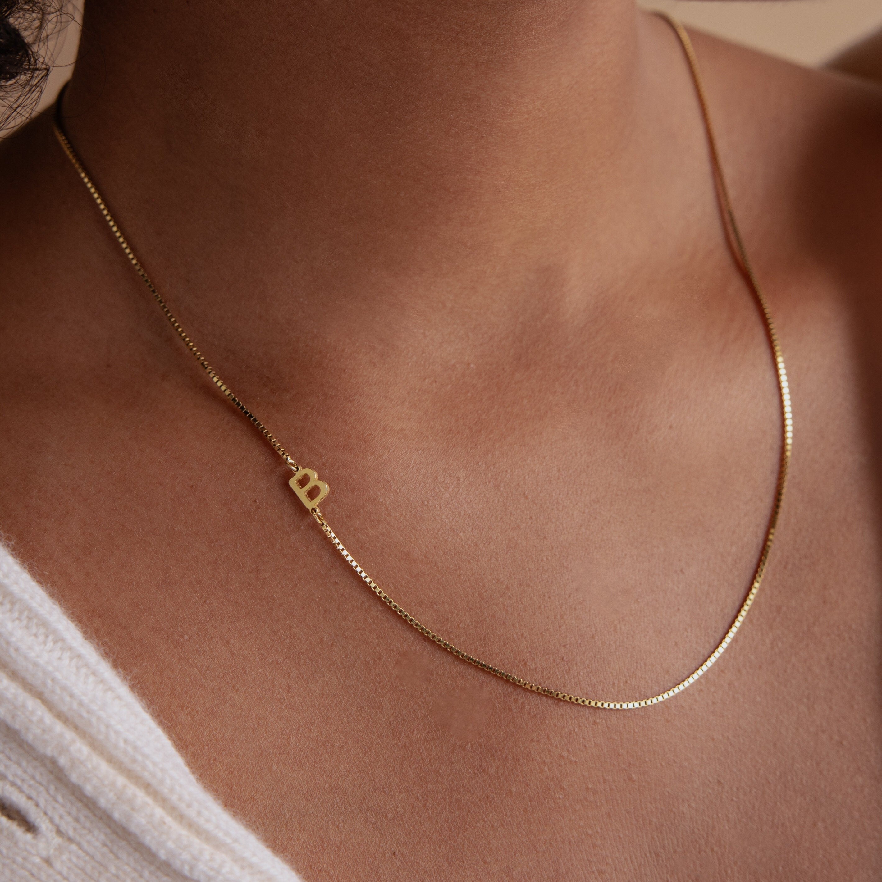 The Sideways Initial Necklace in Box Chain, featuring a small heart charm, rests on the neck and pairs beautifully with a white textured top—an ideal touch of personalized jewelry for everyday elegance.