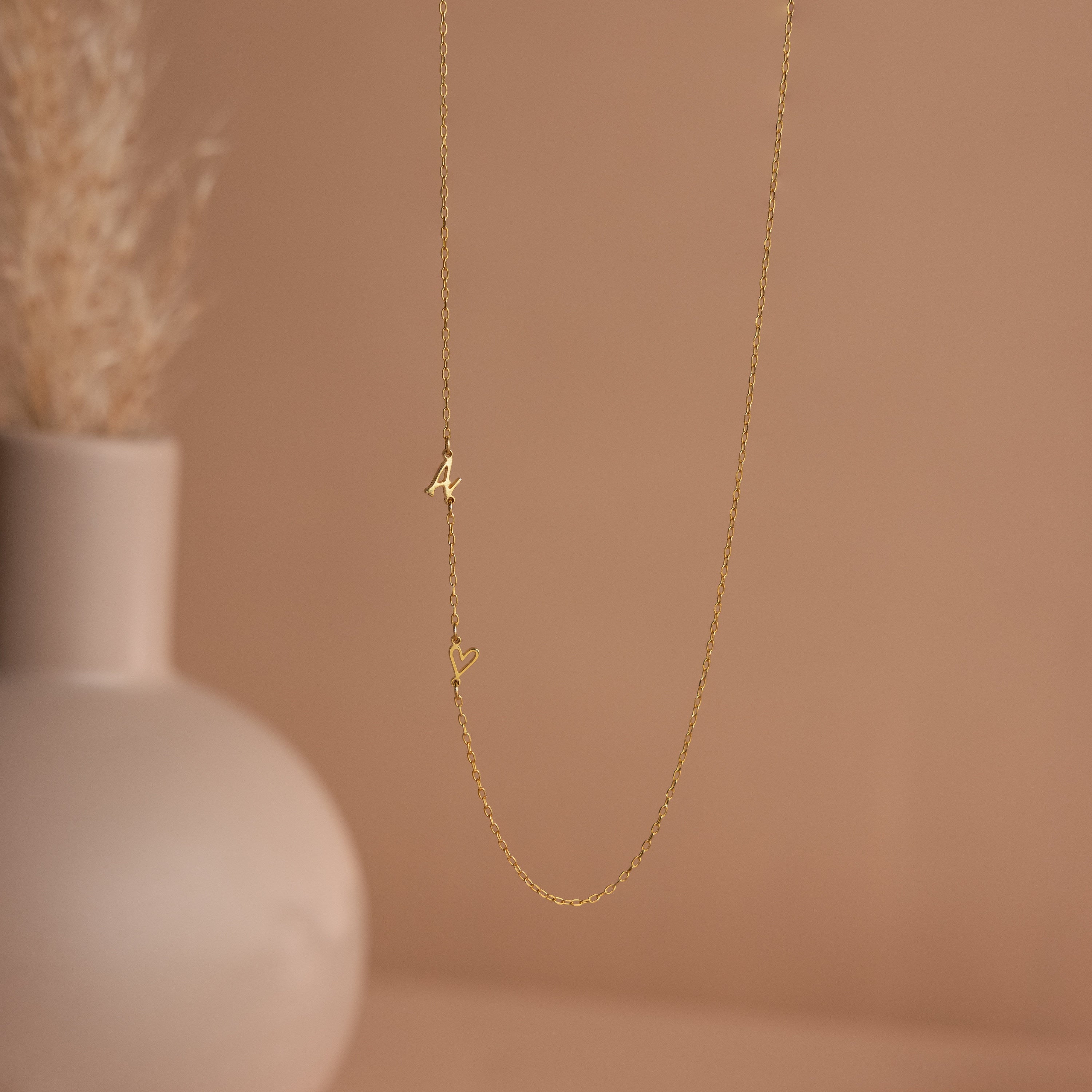 The Sideways Initial Heart Necklace features a letter A and heart pendant, showcased on a beige background with a vase—making it a perfect personalized gift for any occasion.
