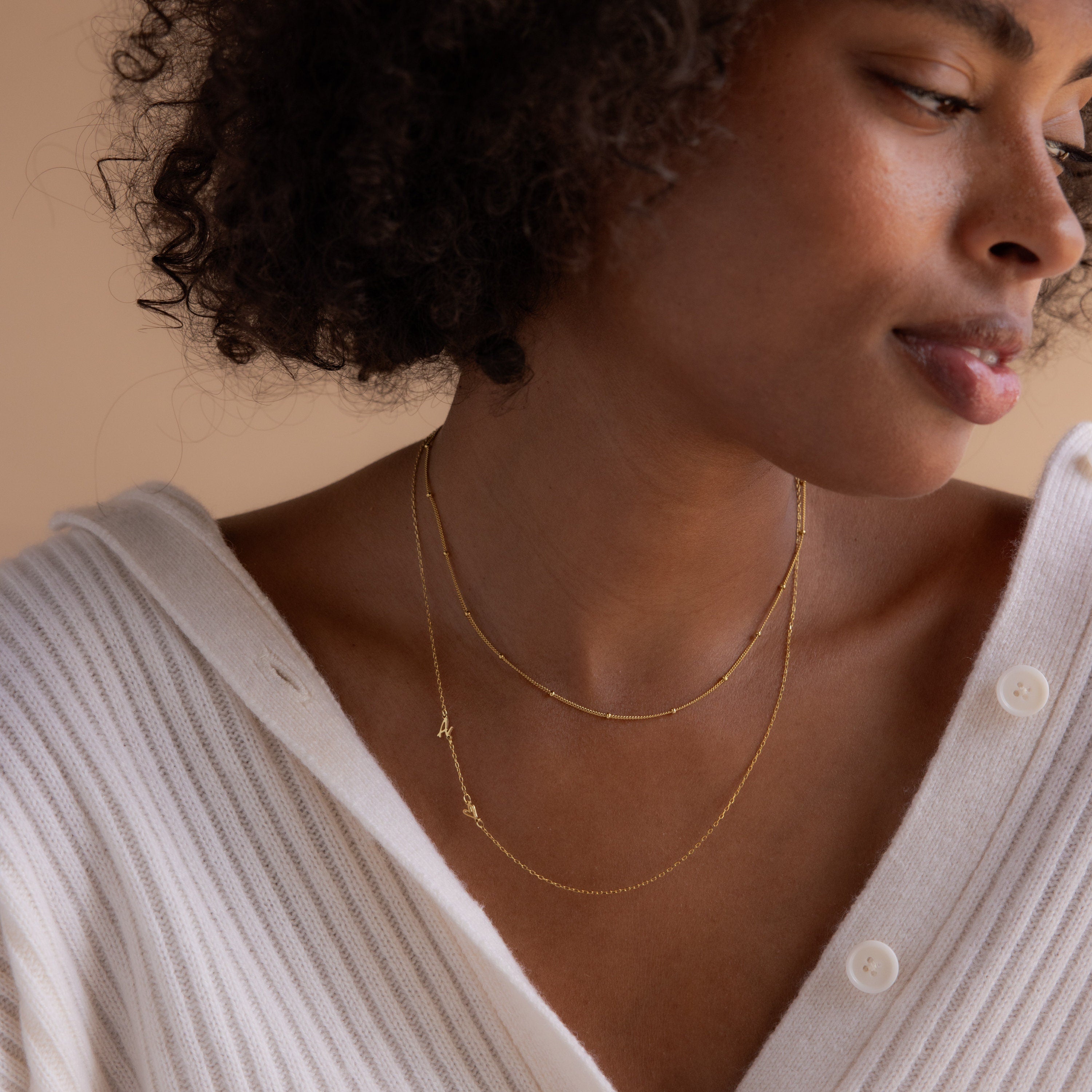 A woman in a white sweater softly smiles as she looks to the side, wearing two delicate gold necklaces, including the Sideways Initial Heart Necklace.