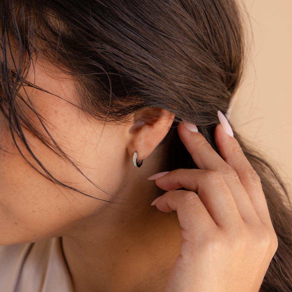 A woman tucks her hair behind her ear, showing off manicured nails and elegant White Opal Inlay Huggies.