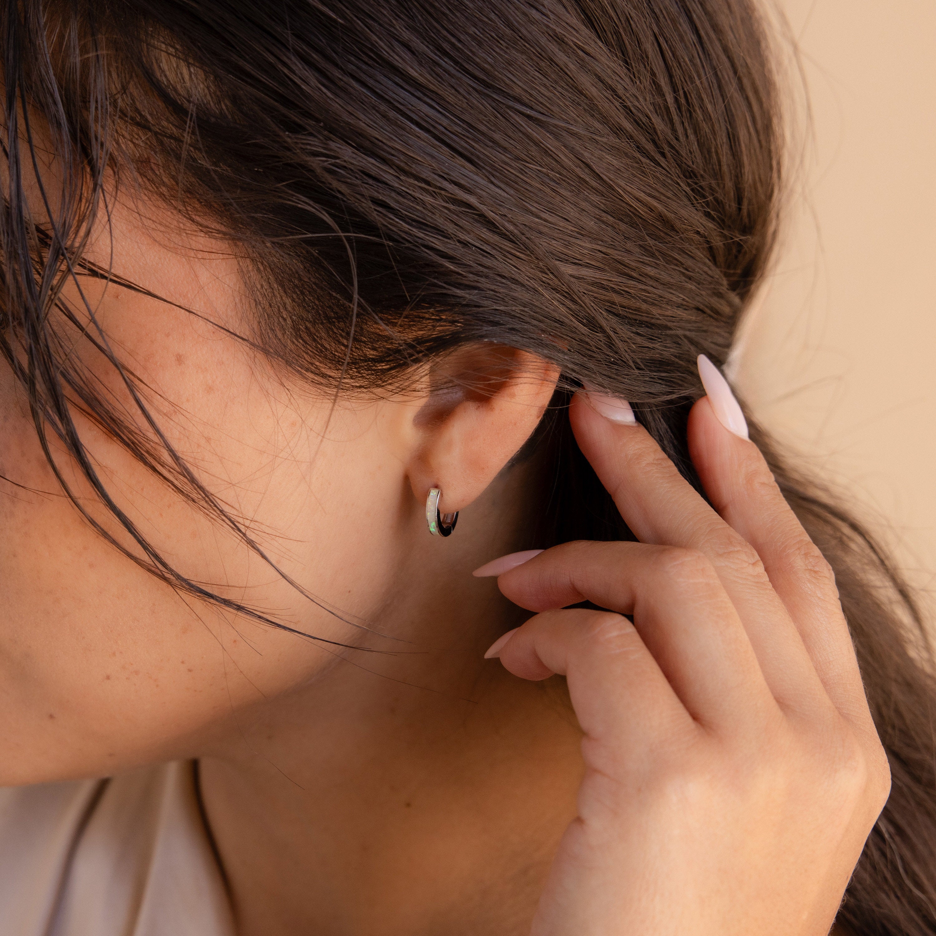 A woman tucks her hair behind her ear, showing off manicured nails and elegant White Opal Inlay Huggies.