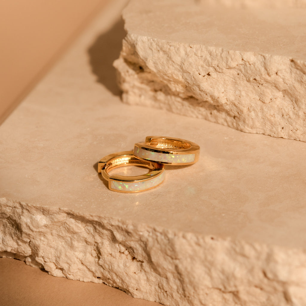 Two White Opal Inlay Huggies adorned with shimmering opals rest on textured stone slabs, glowing warmly under soft lighting.