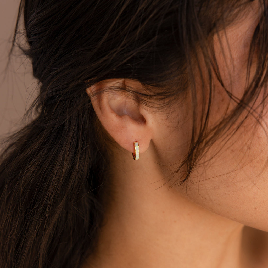 A close-up shows a woman's ear wearing White Opal Inlay Huggies, with her dark hair loosely draped around her face.