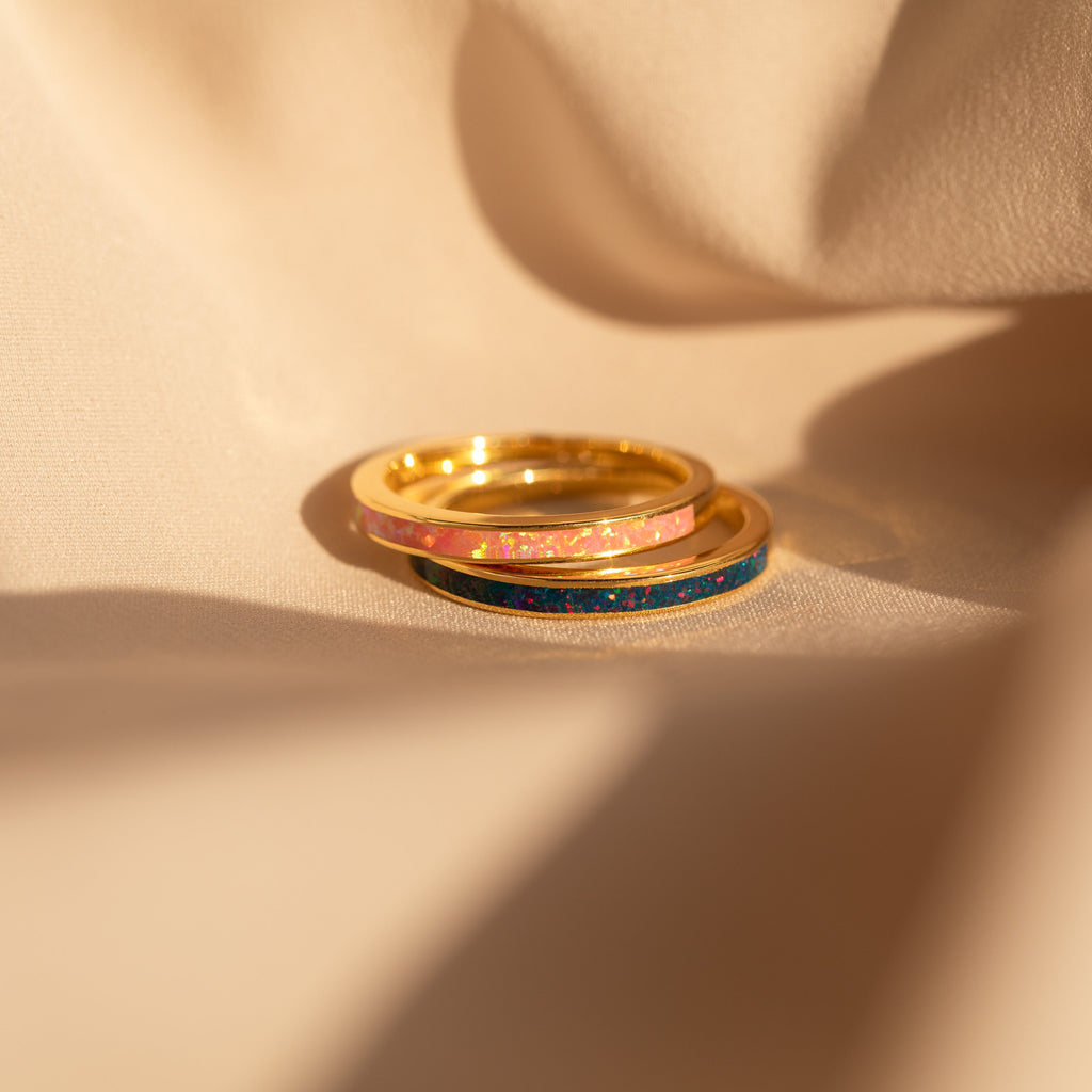 The Pink Opal Inlay Band features a delicate gemstone set in a dainty ring, shown glowing warmly as it rests on soft beige fabric in the sunlight.