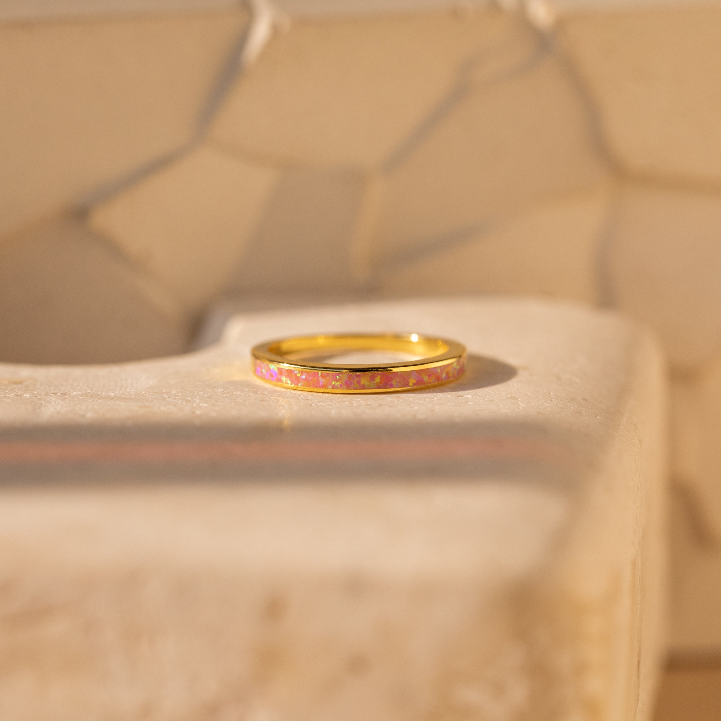 The Pink Opal Inlay Band rests on a textured beige surface, its delicate pink opal inlay glowing softly in the sunlight.