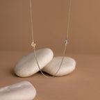 The Birthstone Initial Necklace in Box Chain features an "M" pendant with a small blue gem, elegantly displayed on smooth white stones against a beige background.