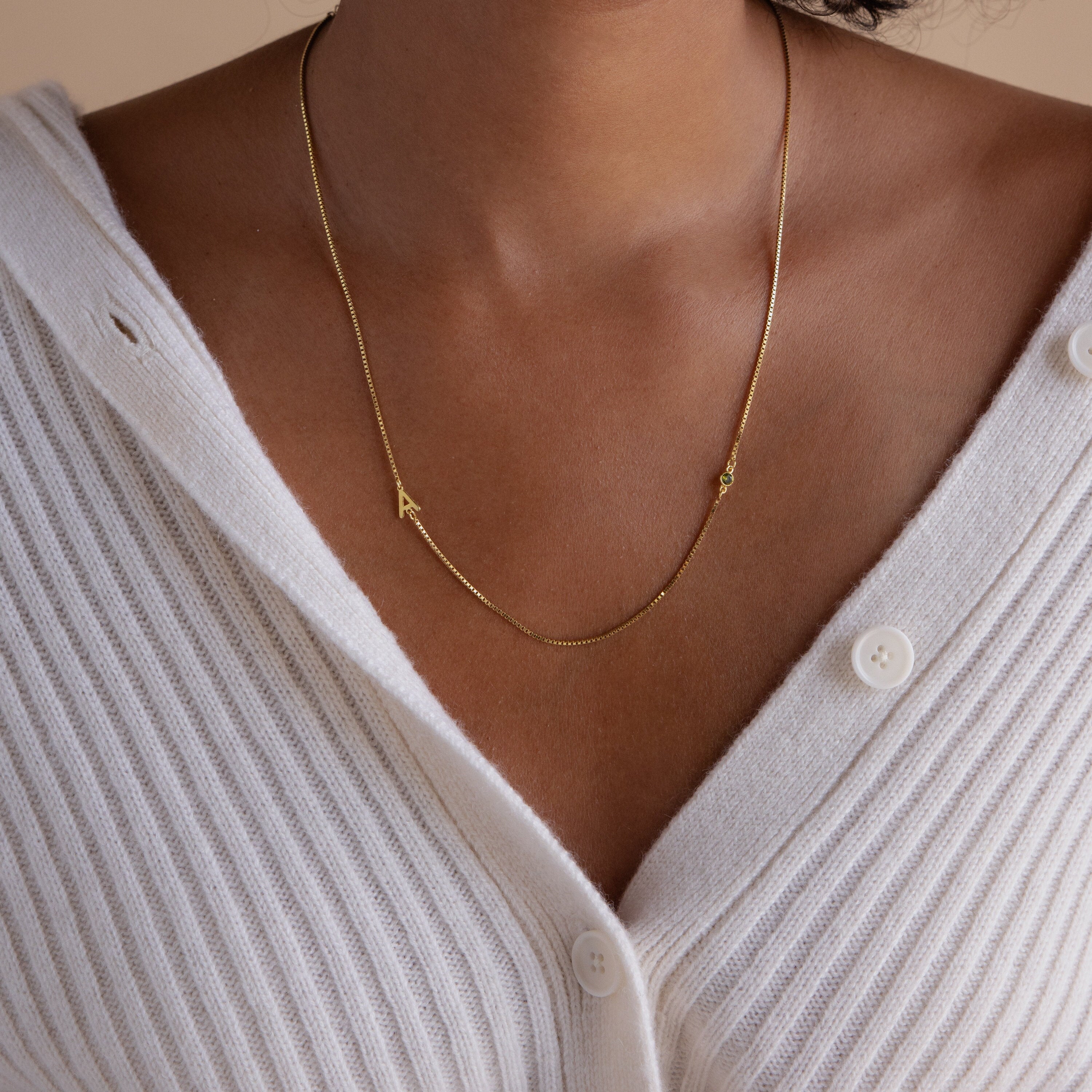 The Birthstone Initial Necklace in Box Chain shines elegantly on a woman wearing a white ribbed buttoned cardigan, personalized with shimmering gold letter charms.