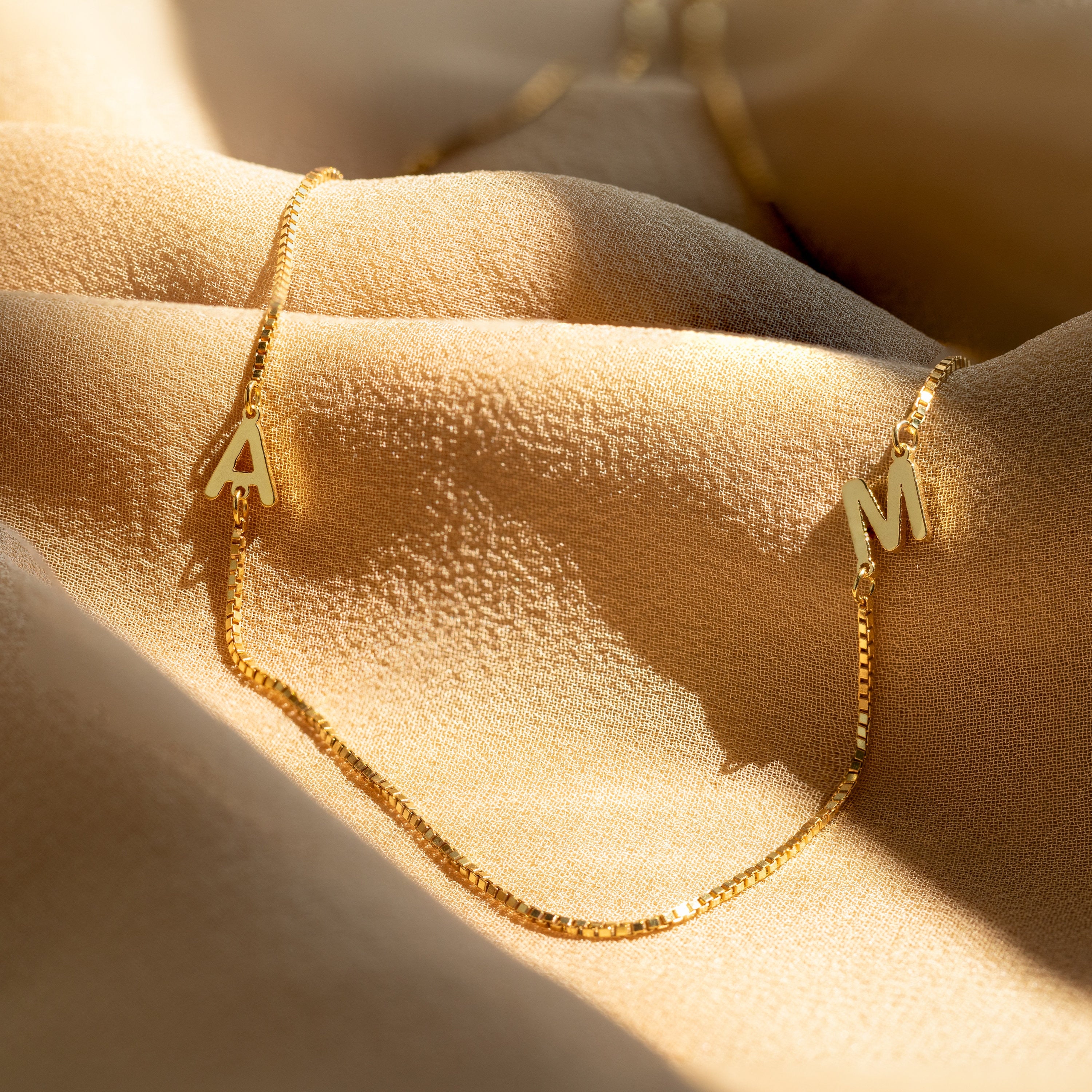 Gold chain bracelet with A and M letter charms on beige fabric, glowing in warm sunlight, pairs perfectly with the Duo Initial Necklace in Box Chain for a personalized charm look.