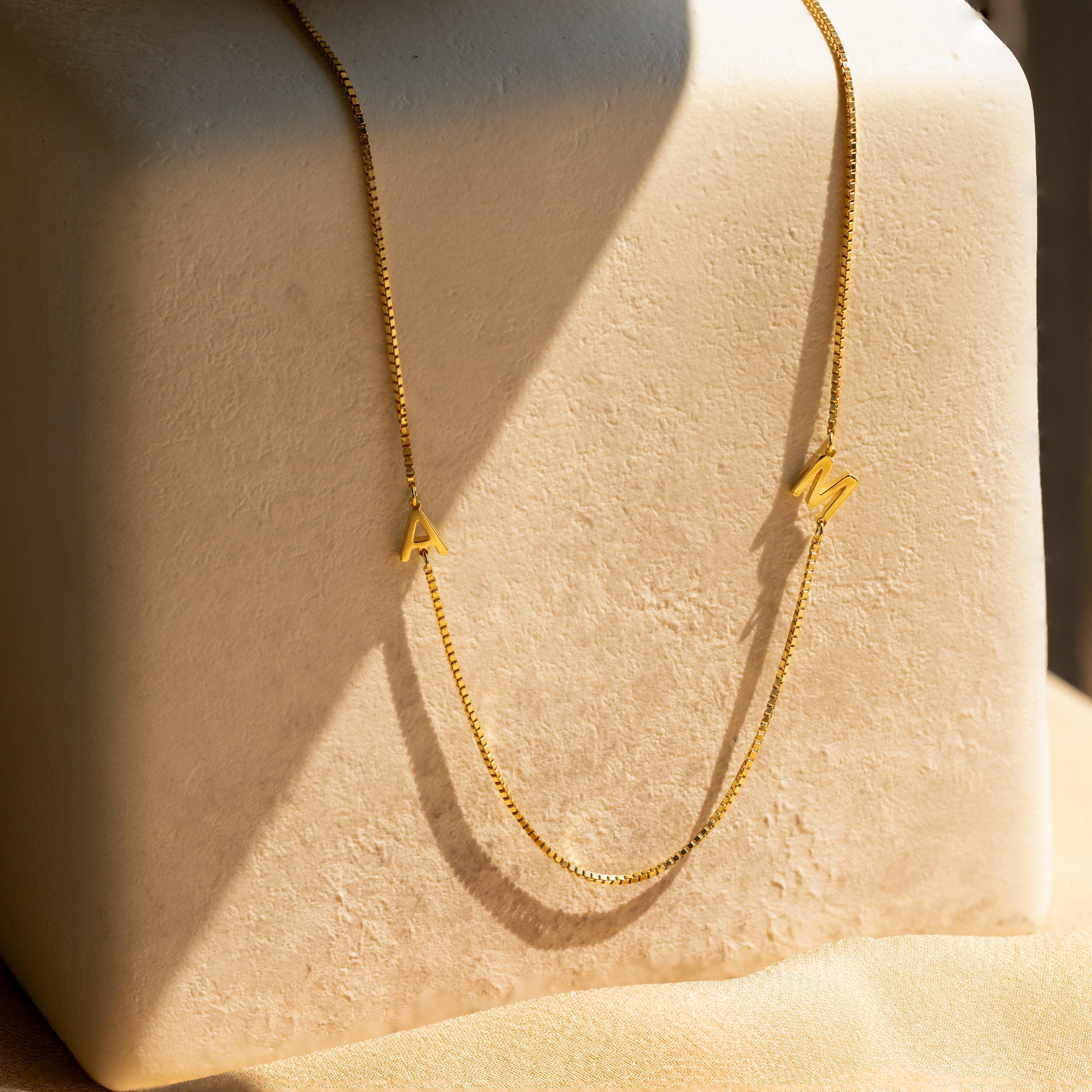 The Duo Initial Necklace in Box Chain features small gold A and M charms, displayed on a textured beige block. This elegant necklace adds a personalized touch to any look.