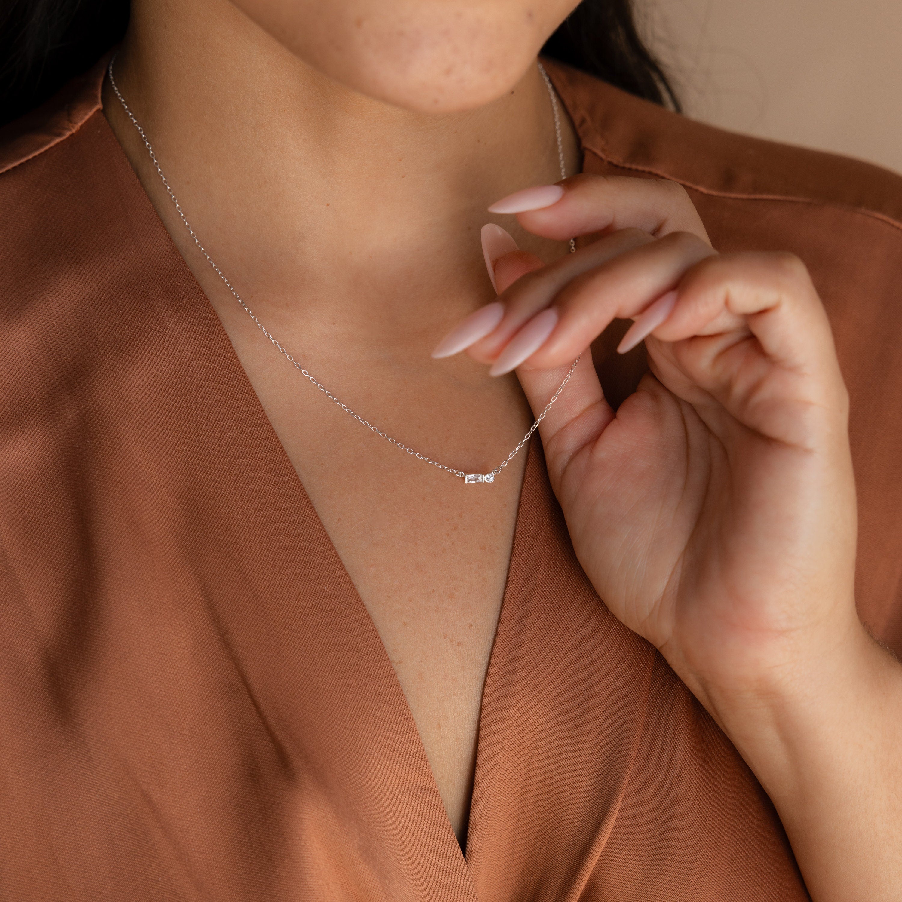 A woman wearing a brown blouse touches an elegant Initial Morse Code Necklace, featuring a delicate silver design with a small pendant.