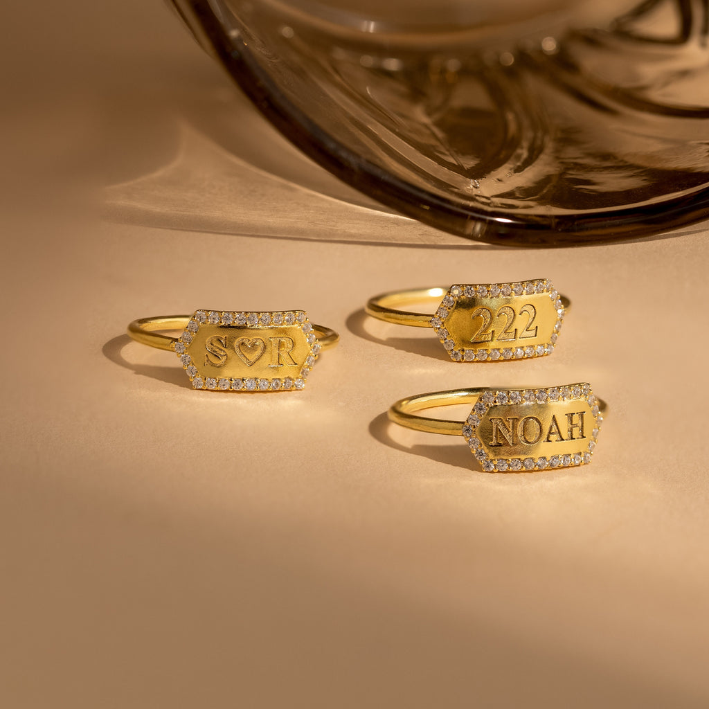 A Custom Pave Hexagon Ring with diamond edges and personalized minimalist engravings—SOR, 222, and NOAH—displayed on a beige surface.