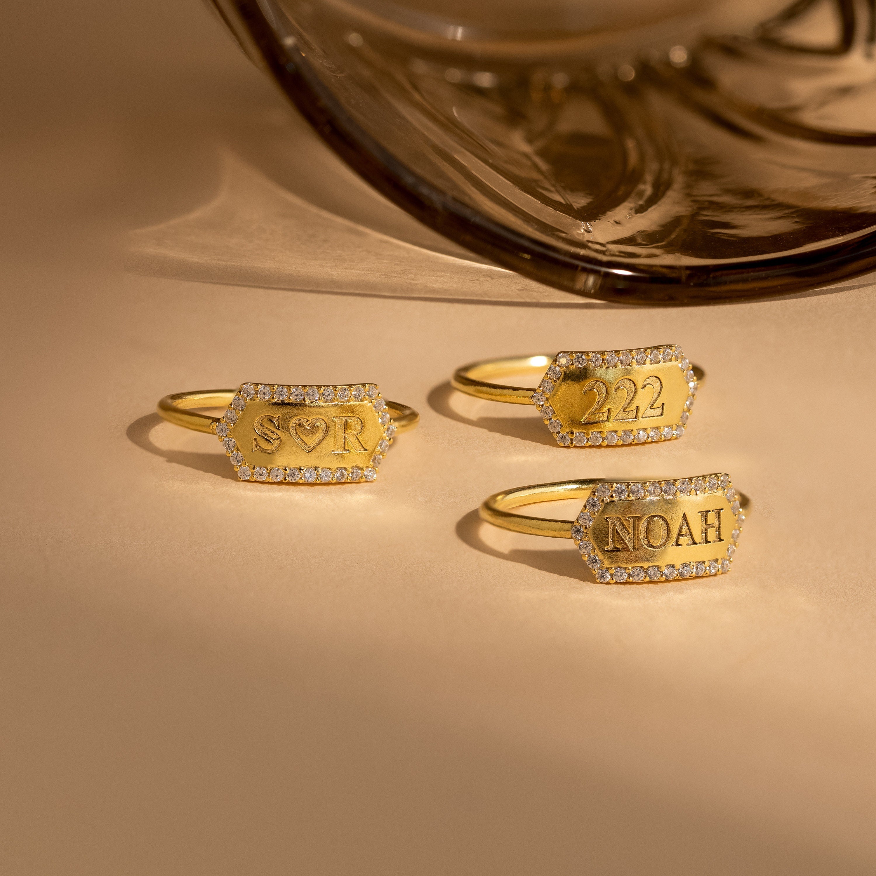 A Custom Pave Hexagon Ring with diamond edges and personalized minimalist engravings—SOR, 222, and NOAH—displayed on a beige surface.