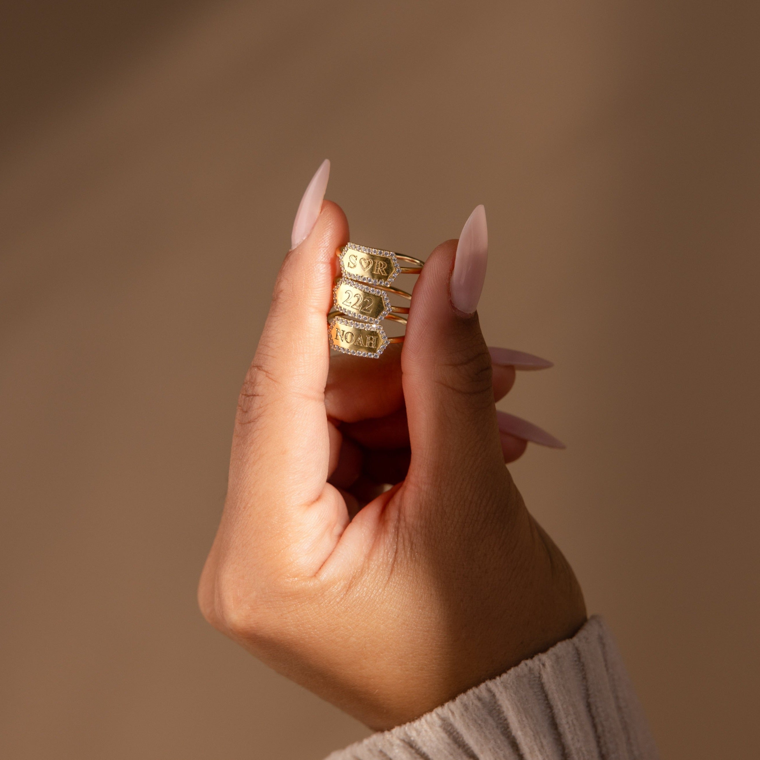 A hand with long nails displays three gold stacking rings, including a personalized minimalist Custom Pave Hexagon Ring, all stacked on one finger against a neutral background.