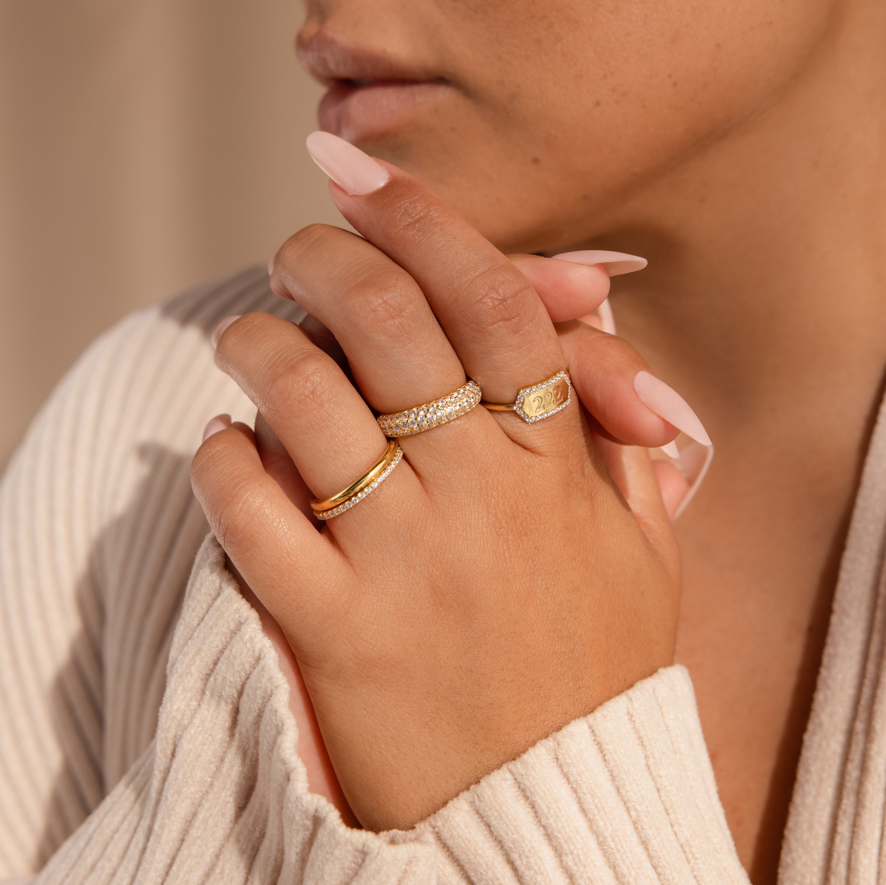 A woman with manicured nails wears multiple gold rings, including an elegant Custom Pave Hexagon Ring, her hands gently clasped near her face.