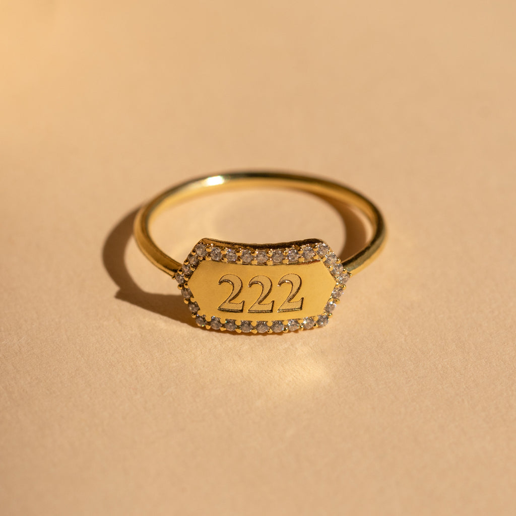 The Custom Pave Hexagon Ring features "222" on a hexagonal face accented with clear stones. This personalized minimalist ring is perfect for stacking and is photographed on a beige background.