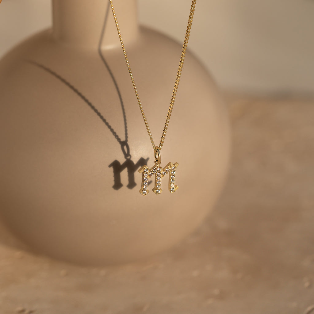 The Gothic Pave Initial Necklace with an M pendant casts a shadow on a round beige vase in soft sunlight—a striking piece of personalized jewelry.