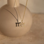 The Gothic Pave Initial Necklace with an M pendant casts a shadow on a round beige vase in soft sunlight—a striking piece of personalized jewelry.