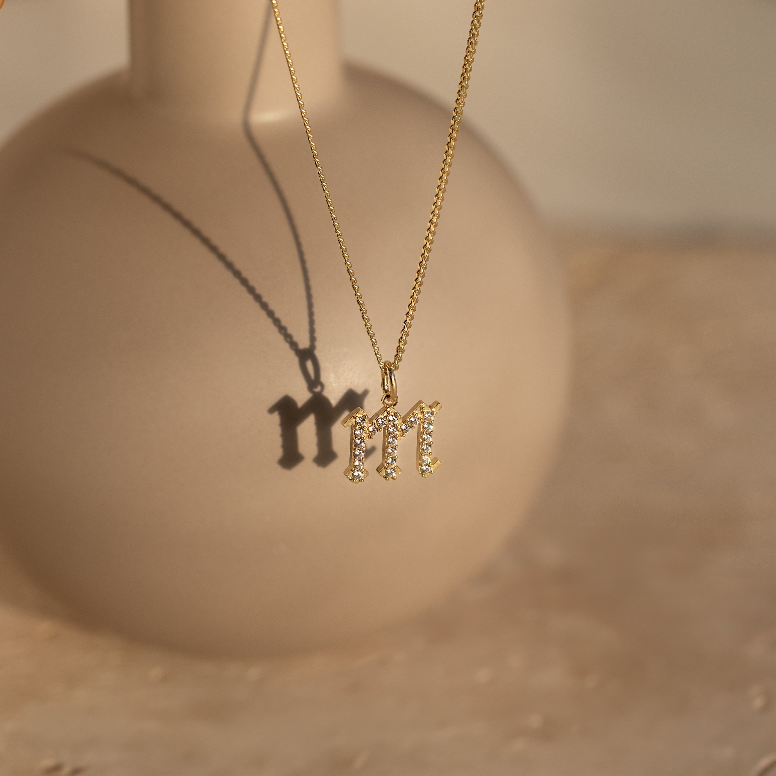 The Gothic Pave Initial Necklace with an M pendant casts a shadow on a round beige vase in soft sunlight—a striking piece of personalized jewelry.