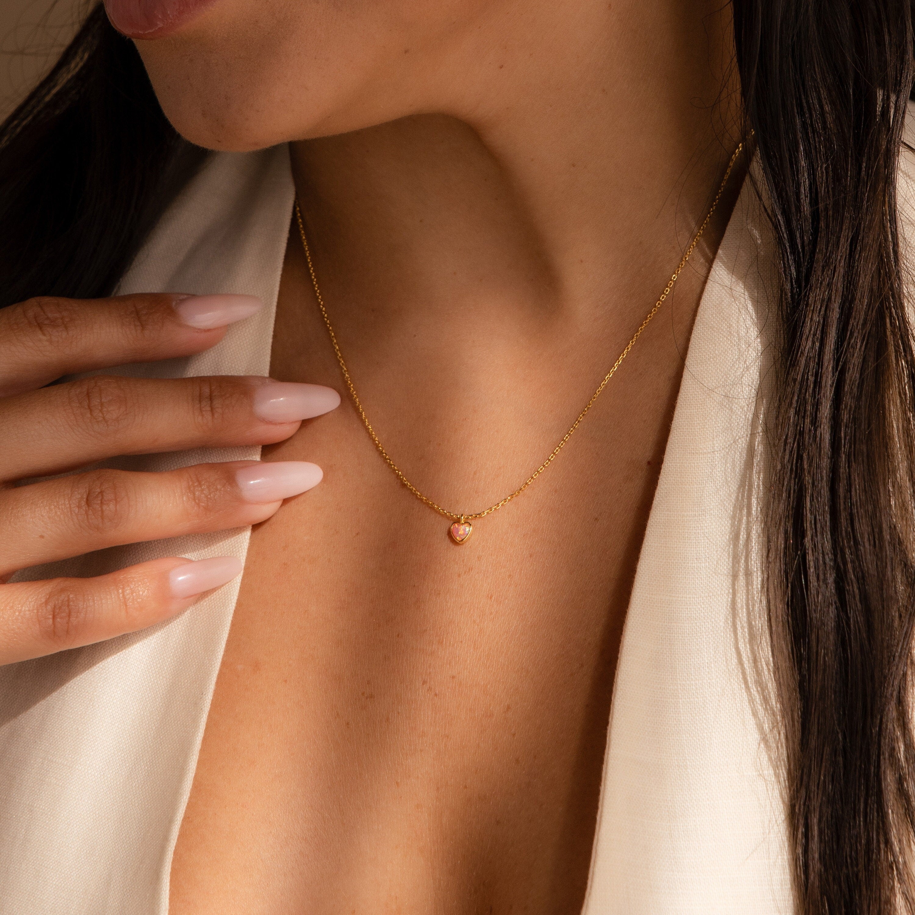 A woman wearing our Pink Opal Heart Necklace, a delicate gold heart-shaped pendant, pairs it with a cream top and gently touches her collarbone with manicured nails.