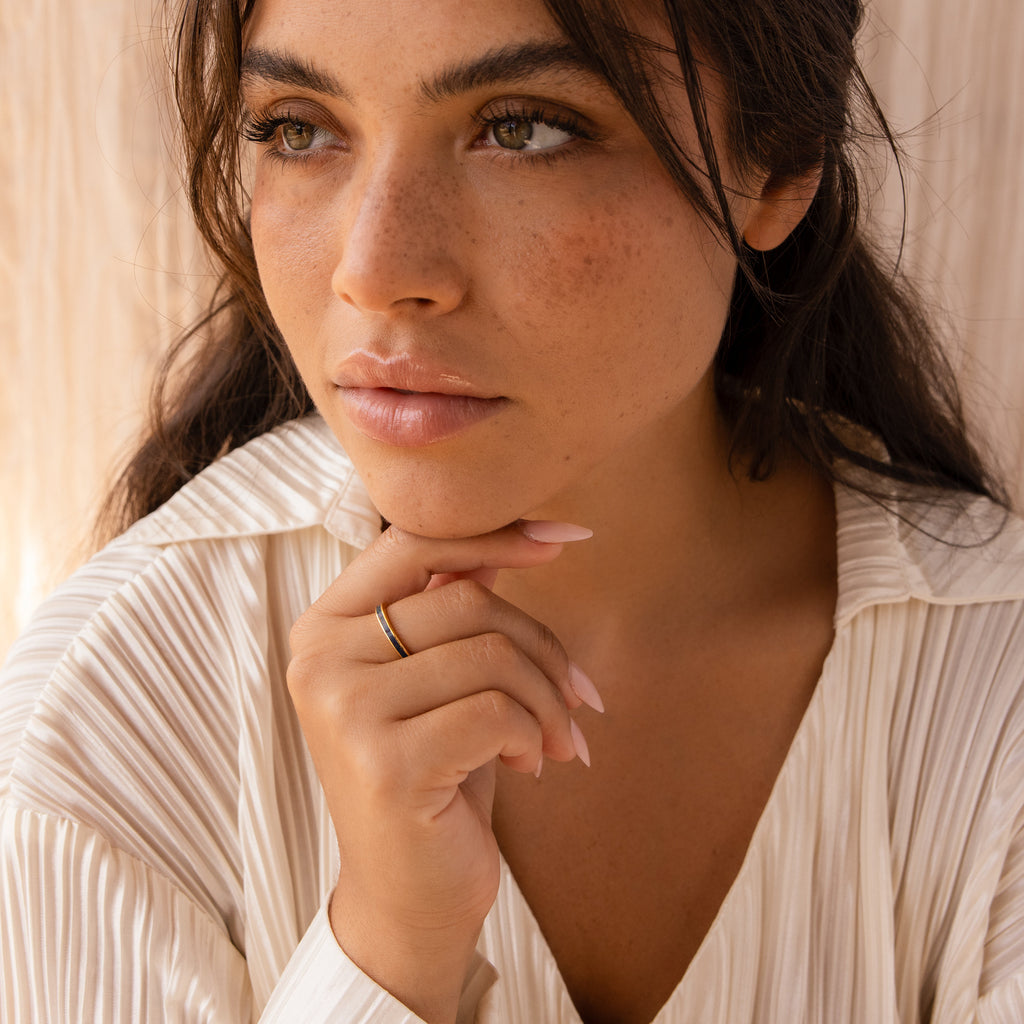 A woman with wavy brown hair and freckles gazes thoughtfully, wearing a cream blouse and a delicate Indigo Opal Inlay Band.
