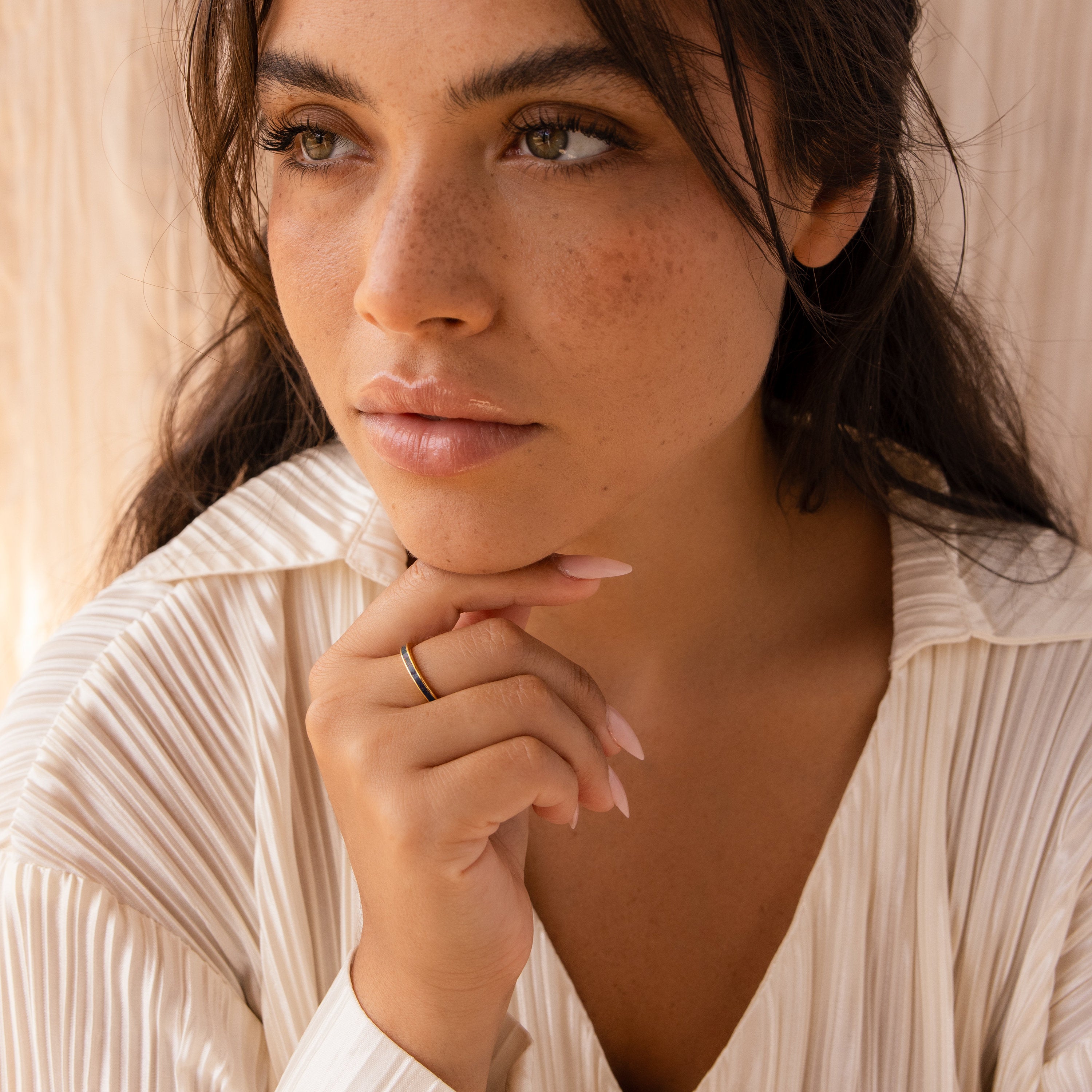 A woman with wavy brown hair and freckles gazes thoughtfully, wearing a cream blouse and a delicate Indigo Opal Inlay Band.