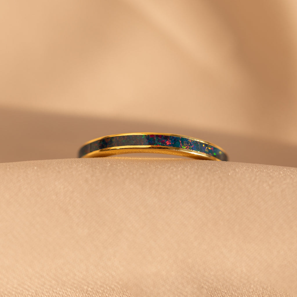 The Indigo Opal Inlay Band, a gold stacking ring featuring a deep, iridescent opal inlay, is displayed on a soft beige fabric background.