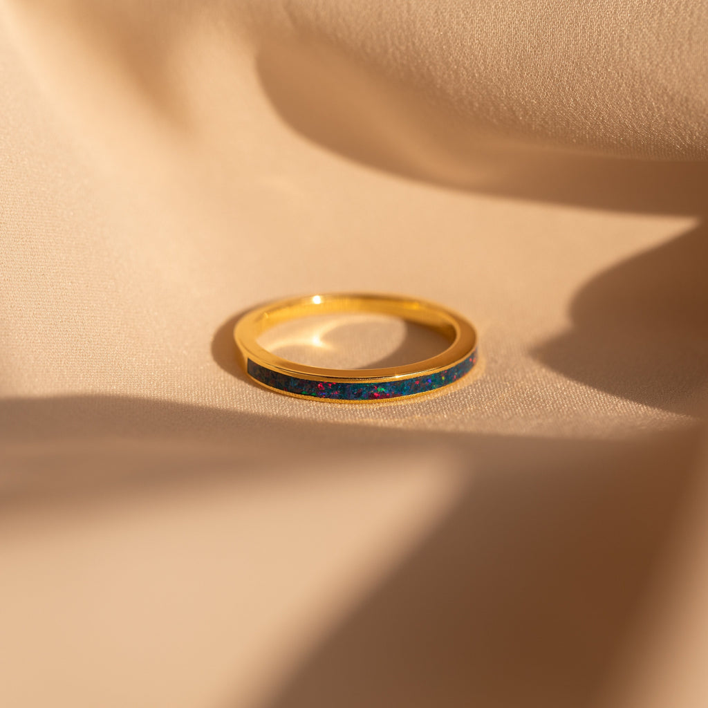 The Indigo Opal Inlay Band, a gold stacking ring featuring a slender blue opal inlay, is displayed on soft beige fabric under warm light.