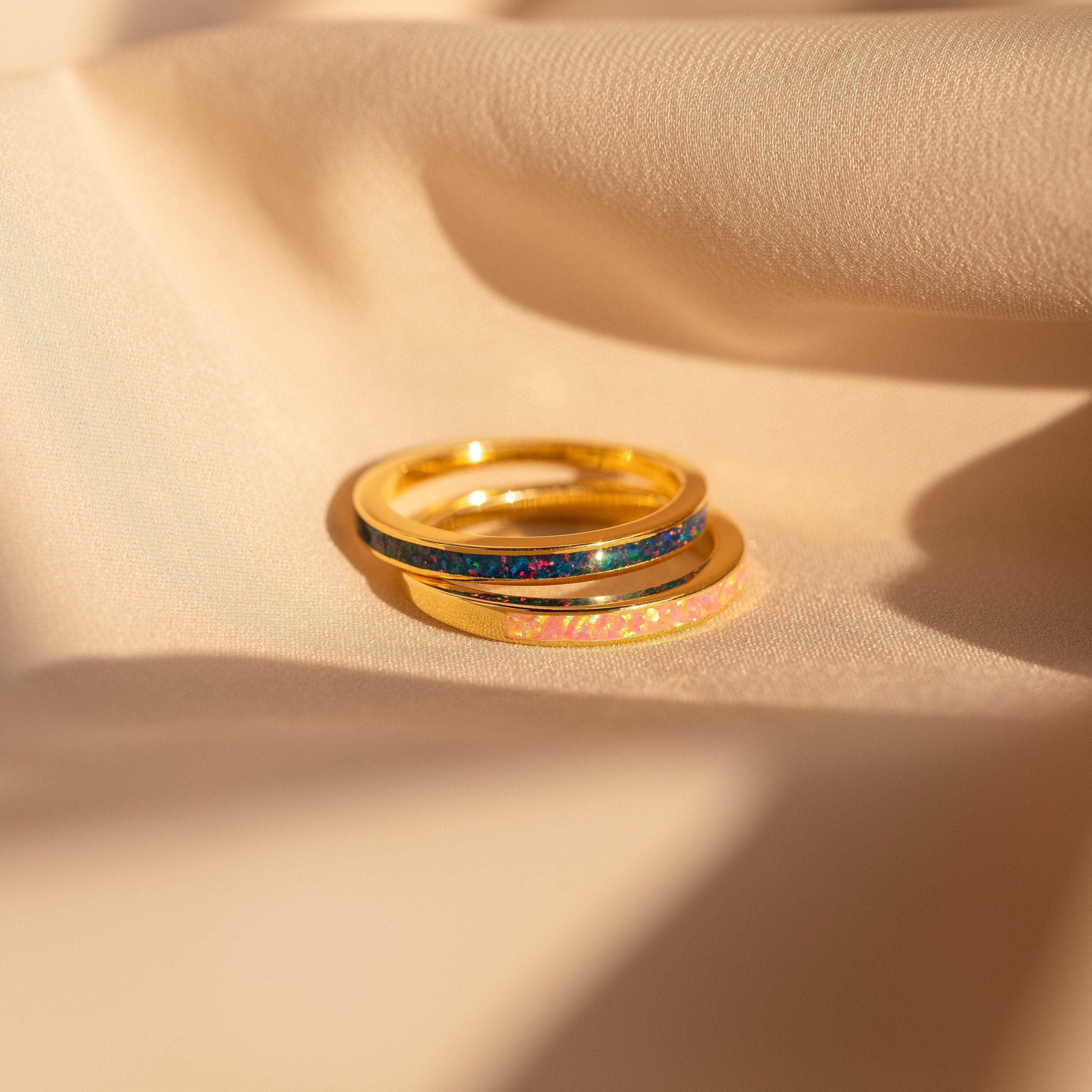 The Indigo Opal Inlay Band and two gold rings are displayed on soft beige fabric, glowing in warm sunlight.