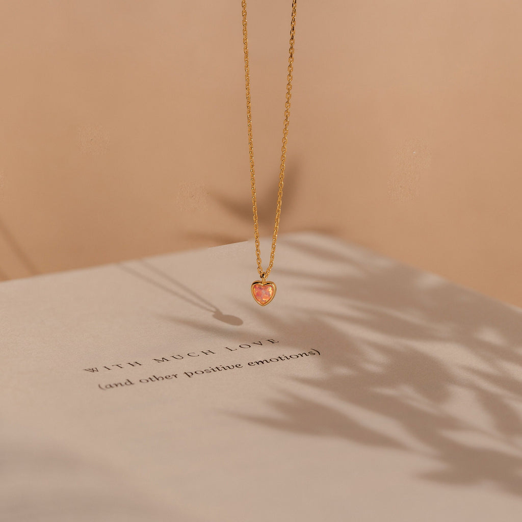 The Pink Opal Heart Necklace, a gold dainty necklace with a heart-shaped pink opal pendant, is displayed resting above a book in soft shadows and warm lighting.