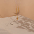 The Pink Opal Heart Necklace, a gold dainty necklace with a heart-shaped pink opal pendant, is displayed resting above a book in soft shadows and warm lighting.
