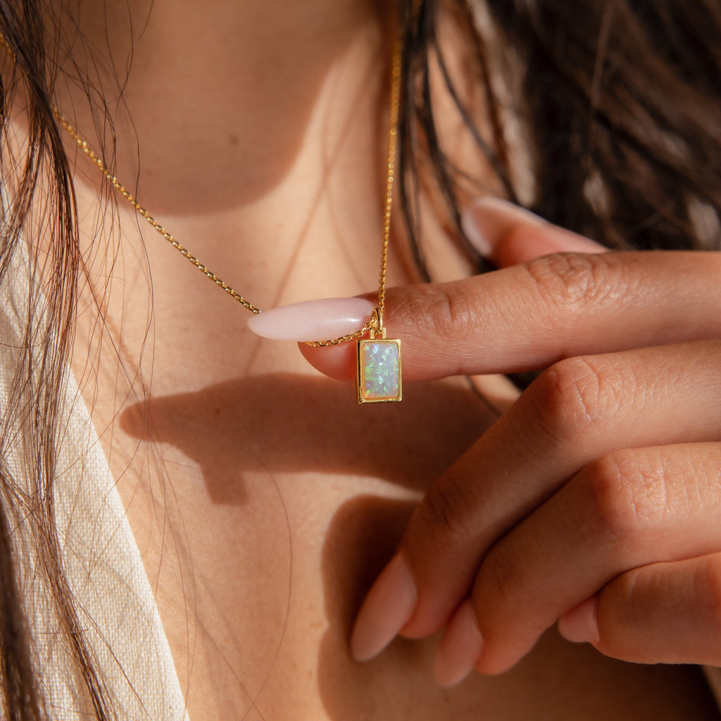 A woman with long nails holds the White Opal Rectangle Necklace with a rectangular pendant near her collarbone, highlighting this elegant and minimalistic piece—ideal as bridal jewelry.