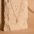 The White Opal Rectangle Necklace features a minimalist design with a white opal charm, perfect for bridal jewelry, shown on a textured beige stone background.