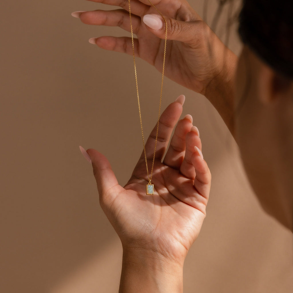 A person holds the White Opal Rectangle Necklace with a rectangular pendant against a beige background—an elegant and minimalistic choice for bridal jewelry.