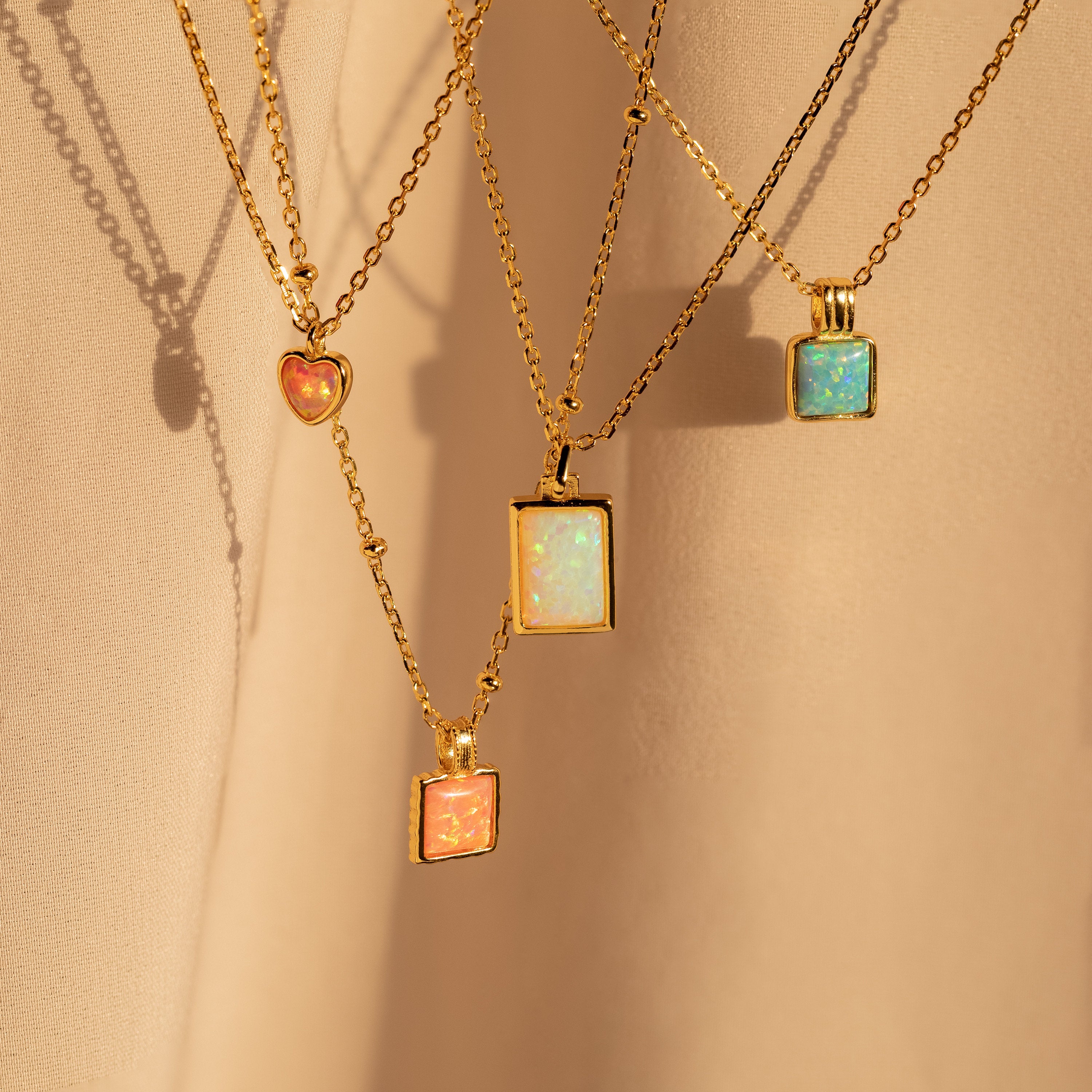 Four gold chain necklaces, including the Pink Opal Pendant Necklace, are displayed on a soft beige fabric background.