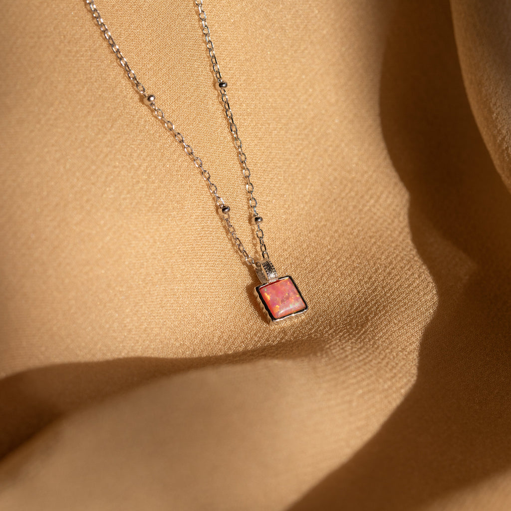 The Pink Opal Pendant Necklace features a square pink opal on a delicate satellite chain, displayed on a beige textured fabric background for an elegant, minimalist look.
