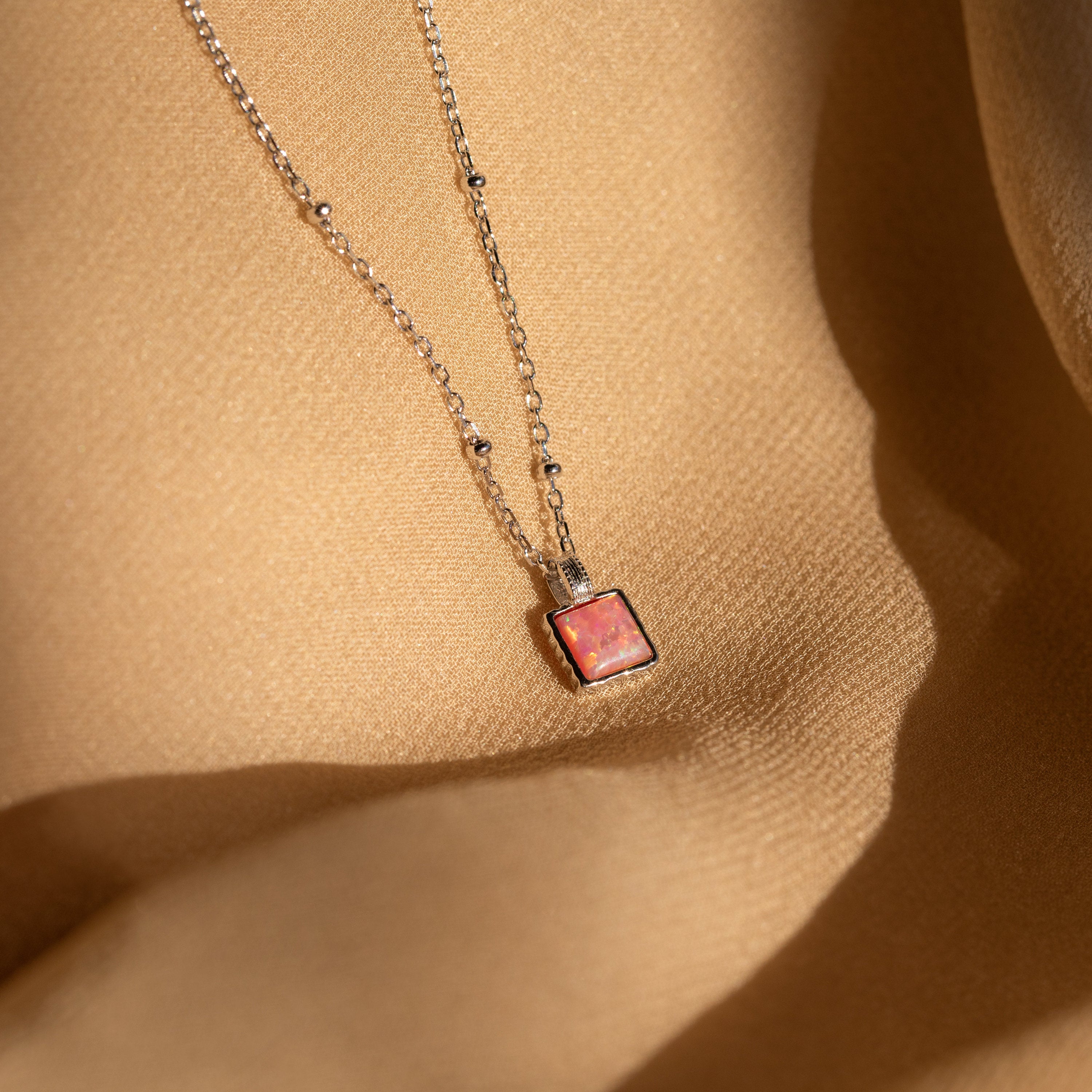 The Pink Opal Pendant Necklace features a square pink opal on a delicate satellite chain, displayed on a beige textured fabric background for an elegant, minimalist look.