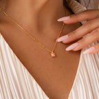 Gold necklace with a square pink opal pendant, worn by a woman in a pleated white top with manicured nails touching the chain.