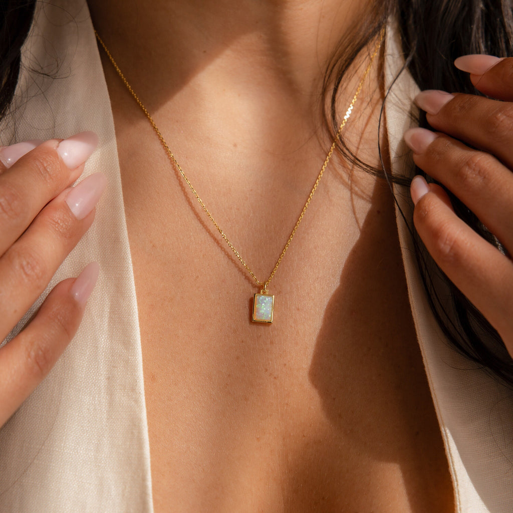 Gold necklace with a rectangular opal pendant, worn by a woman in a light top with hands framing the jewelry.
