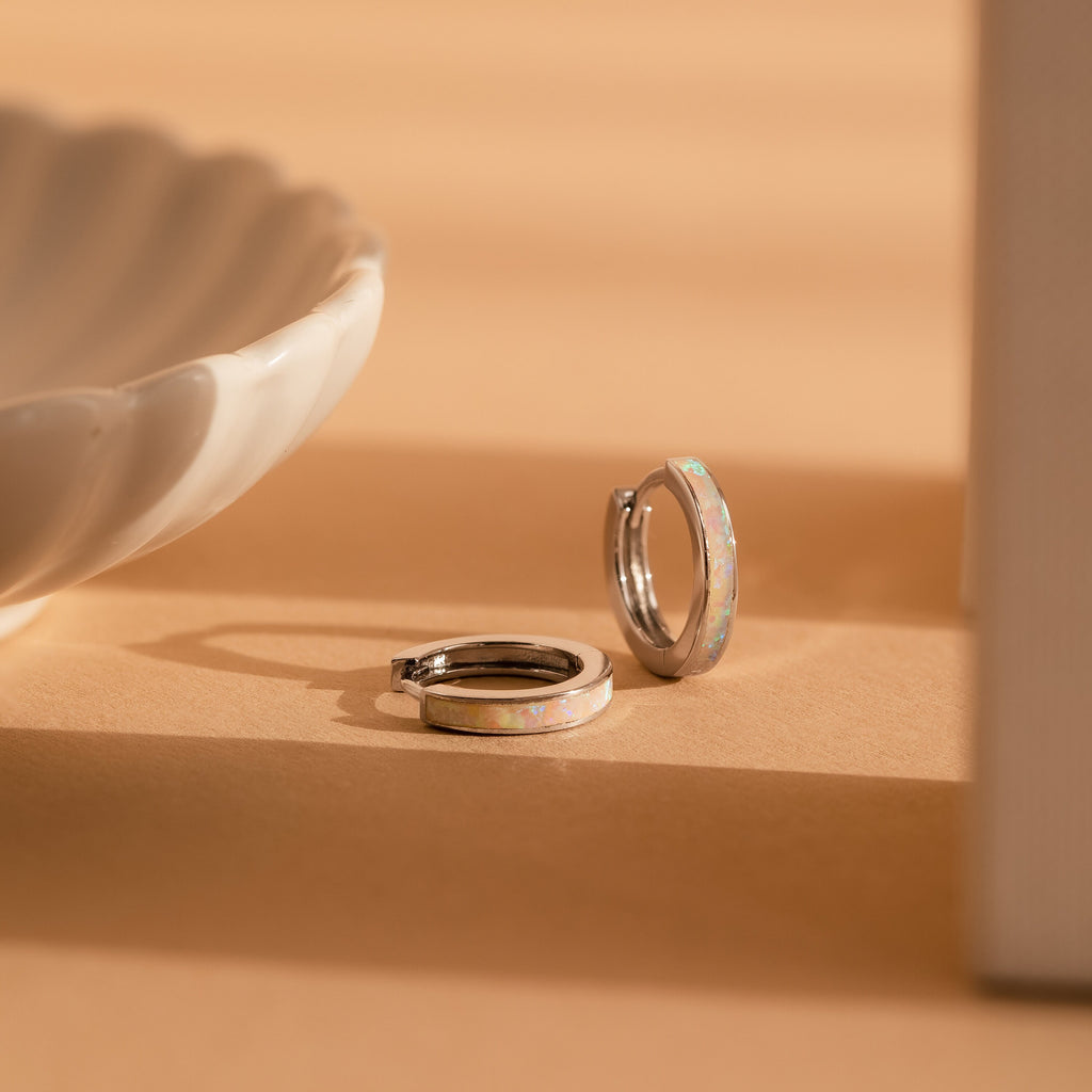 White Opal Inlay Hoops featuring shimmering opal accents rest on a beige surface beside a white ceramic dish—ideal as elegant bridal earrings or for everyday wear.