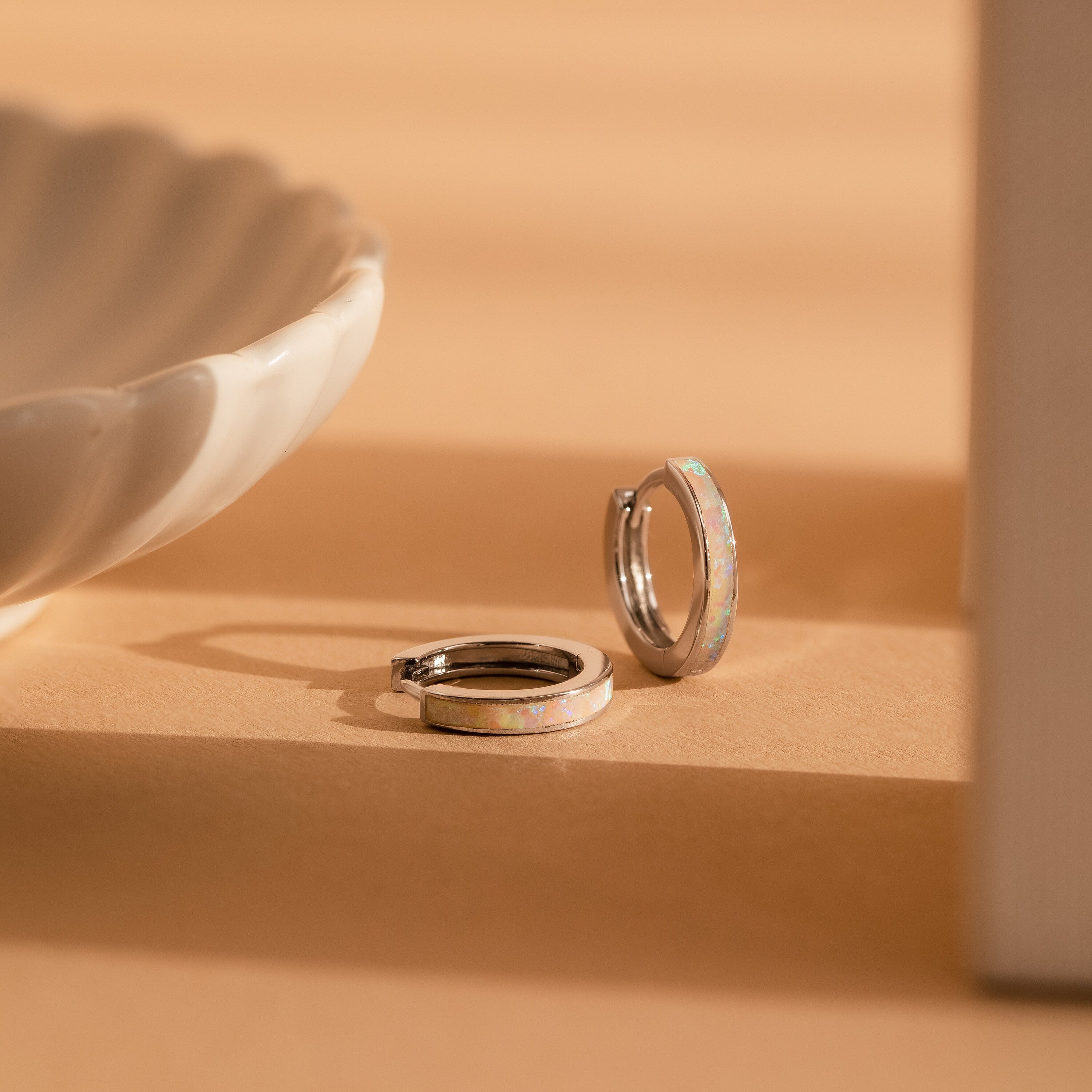 White Opal Inlay Hoops featuring shimmering opal accents rest on a beige surface beside a white ceramic dish—ideal as elegant bridal earrings or for everyday wear.