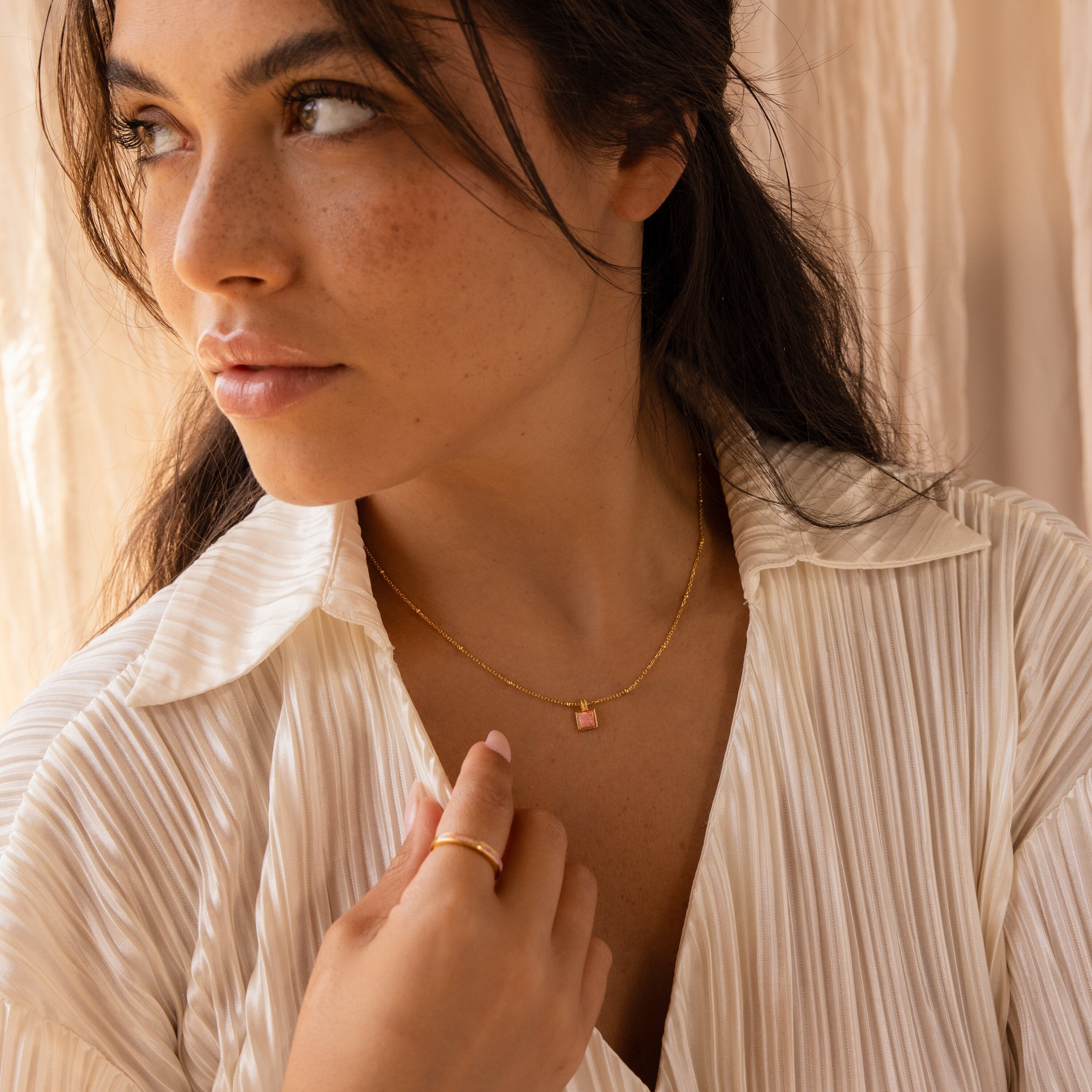 Woman in a cream pleated blouse wears the Pink Opal Pendant Necklace and a gold ring, gazing thoughtfully to the side.