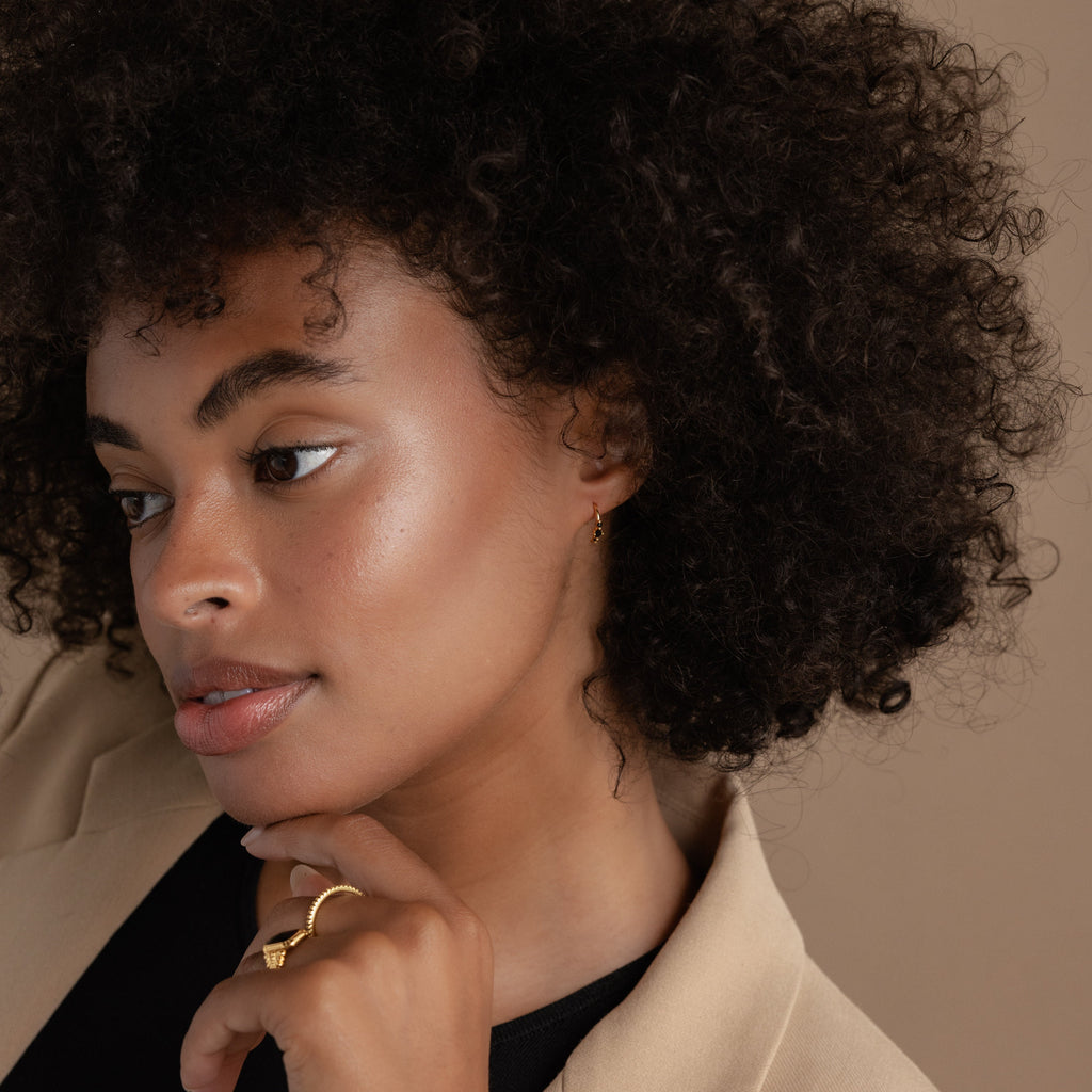 Woman with curly hair in a beige blazer, looking to the side with hand near her chin, wears gold pieces from the latest Jewelry Collection and striking Amaya Black Marquise Threaders.